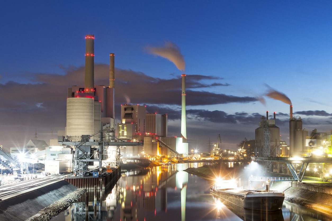 A coal power plant sits at the river's edge at dusk.  Lights from the buildings and streets glow, and a reflection of the whole scene can be seen on the water of the calm river below.